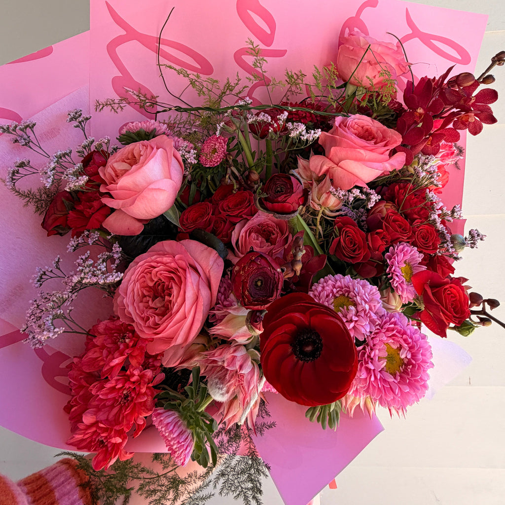 A vibrant floral bouquet featuring pink and red roses, ranunculus, and hydrangeas wrapped in bright pink patterned paper, held by a hand wearing a striped sweater against a white paneled wall.