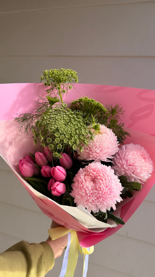 A bouquet of pink flowers wrapped in pink paper, held by a hand against a light-colored paneled wall.