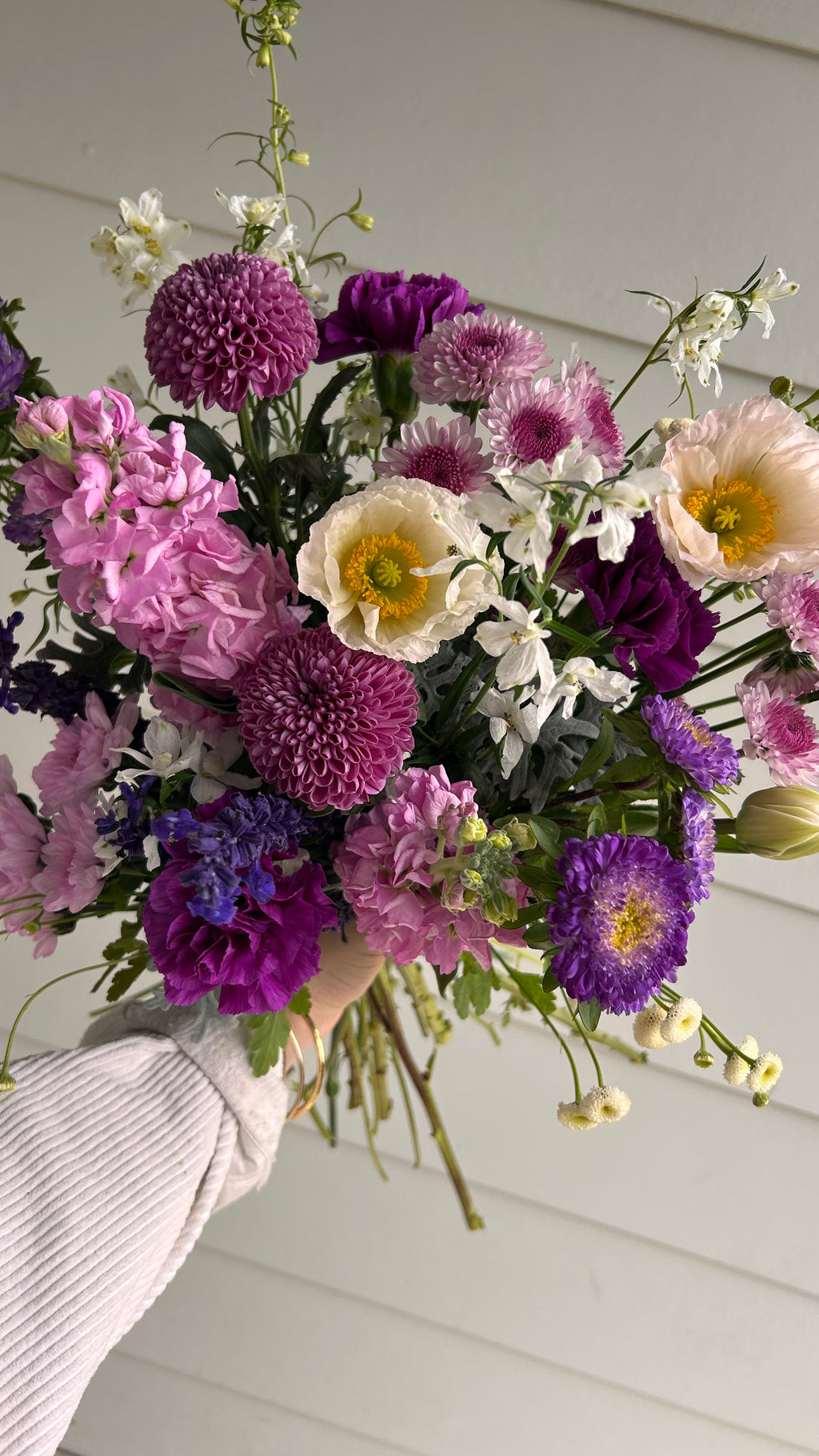 A hand holding a vibrant mixed flower bouquet featuring purple and pink chrysanthemums, white daisies, and green foliage, set against a white paneled wall.