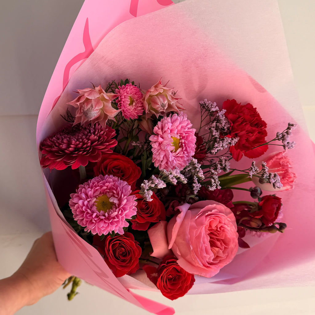 A bouquet of pink and red flowers wrapped in pink tissue paper, held by a hand against a plain light background.